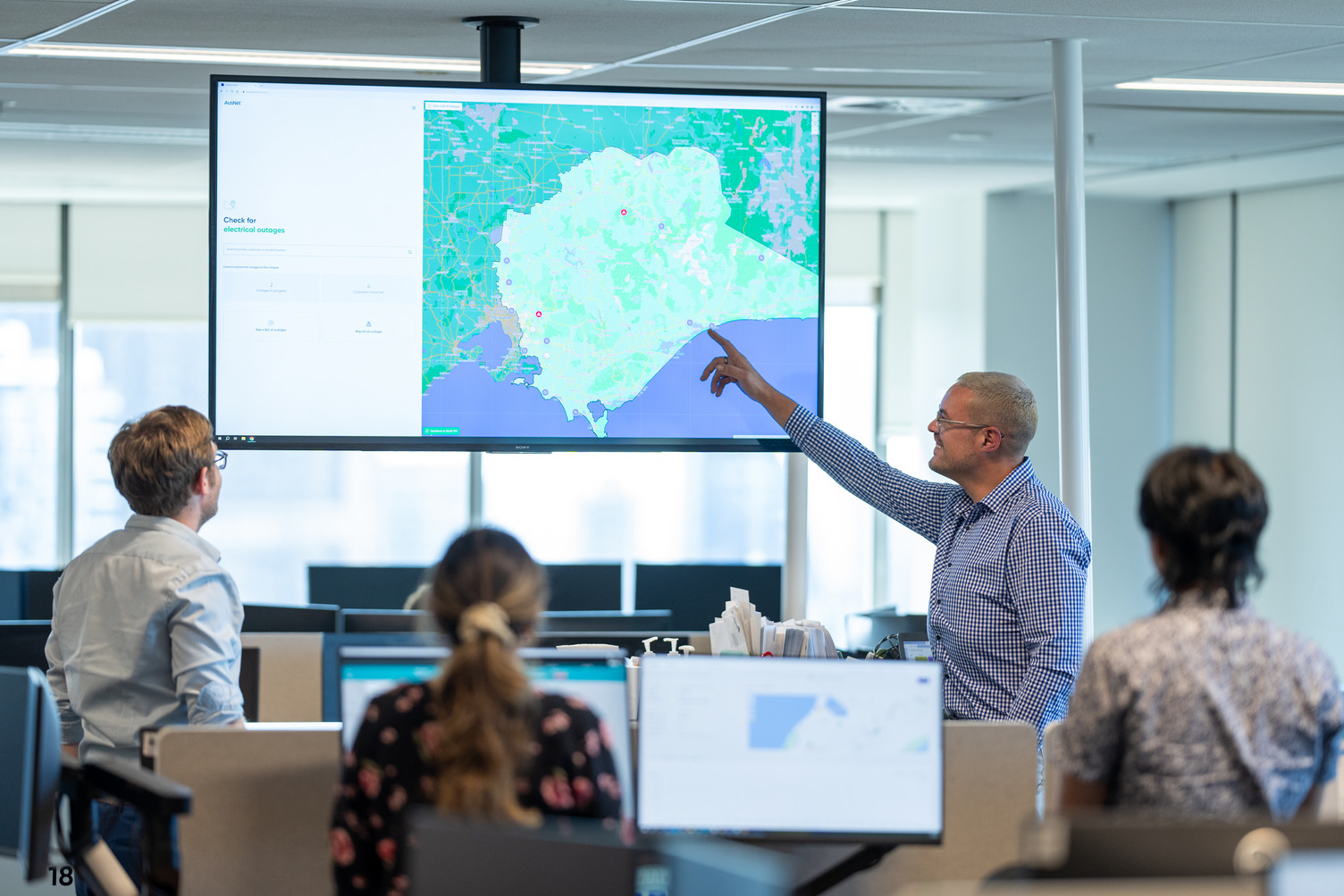 A man in an office presenting to other people while pointing at a TV