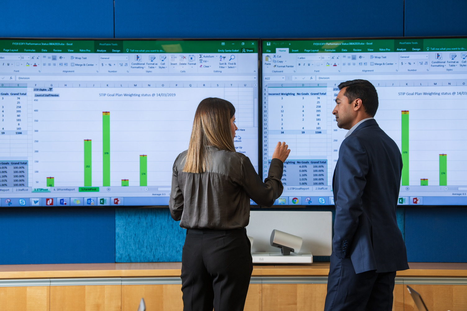 A man and woman talking infront of a screen showing graphs and charts